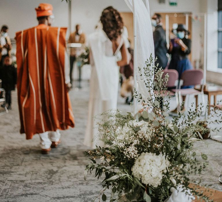 White frame wedding arch with draping, mixed white flowers and green foliage with bride and guest in traditional orange and white outfit in the background at Bridge Community Church Wedding