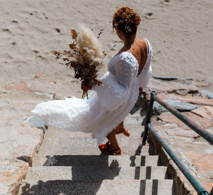 Boho bride walks down beach steps holding bouquet