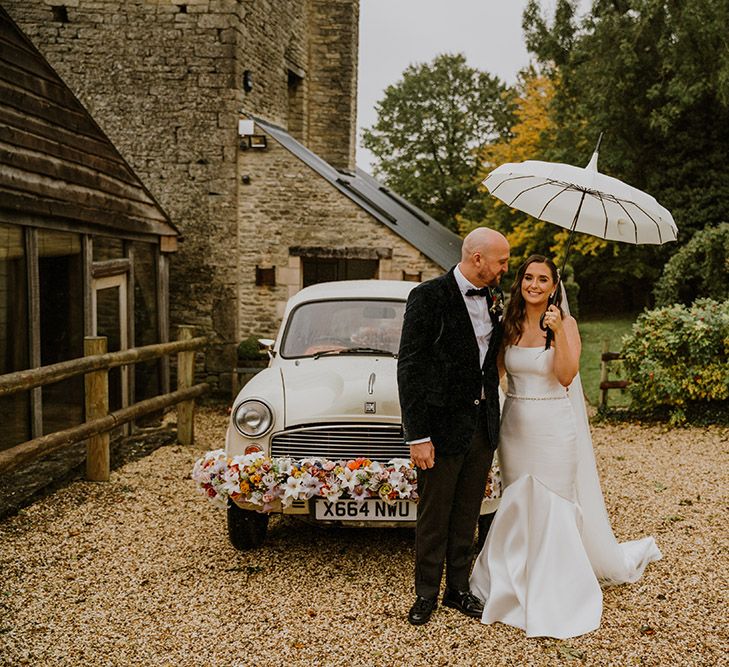 Bride in a strapless wedding dress with mermaid tail holding an umbrella stood next to her groom in a velvet tuxedo and their wedding car with floral bumper decoration 