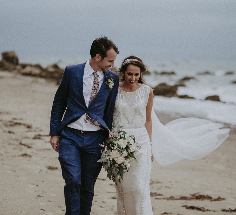 Bride & groom walk along the beach together with the sea in the background