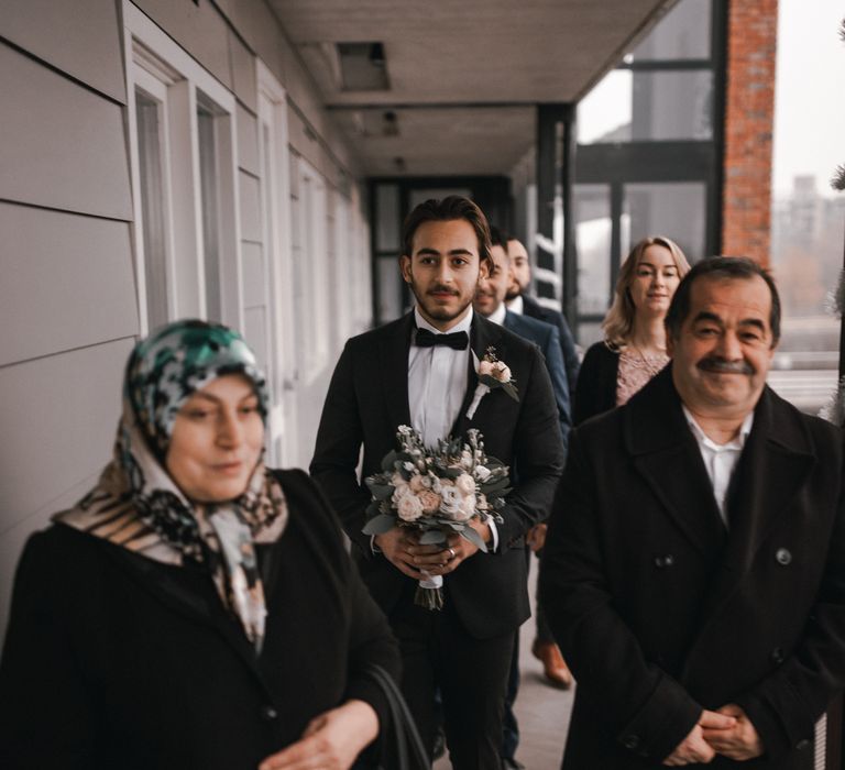 Groom walks with wedding party as he carries floral bouquet