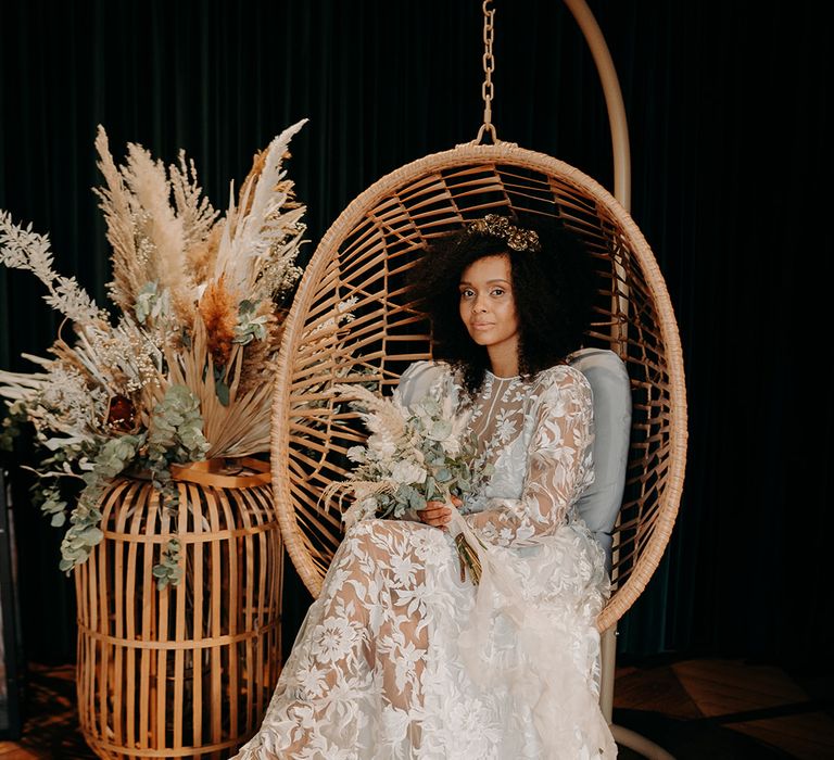 Bride sits in wicker chair surrounded by pampas grass and holding floral bouquet