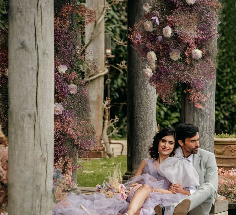 Bride & groom sit beside lake as bride leans into groom