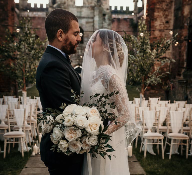 Bride and groom embracing at the altar with the bride wearing a sheer veil over her face and holding a white rose wedding bouquet 