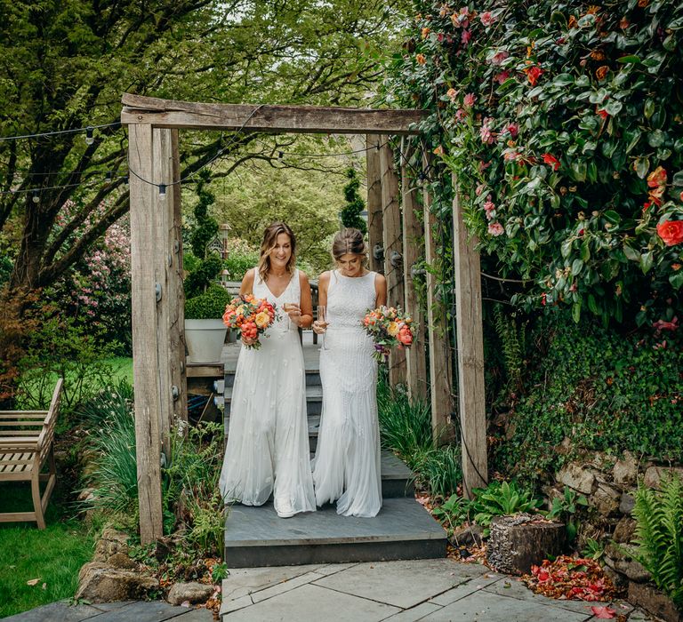 Same-sex couple walk together through gardens at Ever After wedding venue whilst holding bright floral bouquets 