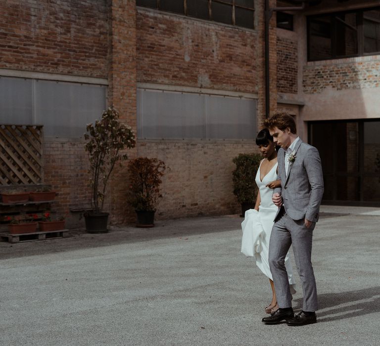 A bride and groom walk outside their venue. He wears a grey suit and she holds up the skirt of her long white dress.