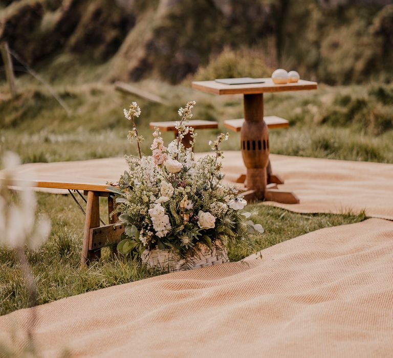 Pink, green and white mixed floral arrangement in white woven basket by wooden bench at clifftop ceremony for Dunluce Castle wedding