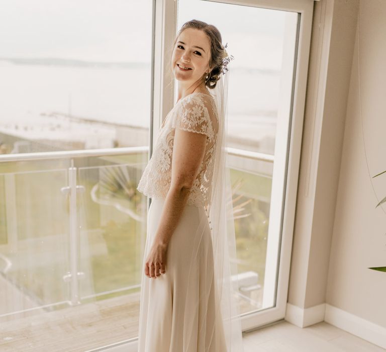 Bride in full-length lace top wedding dress with capped sleeves and veil standing by window before Dunluce Castle wedding