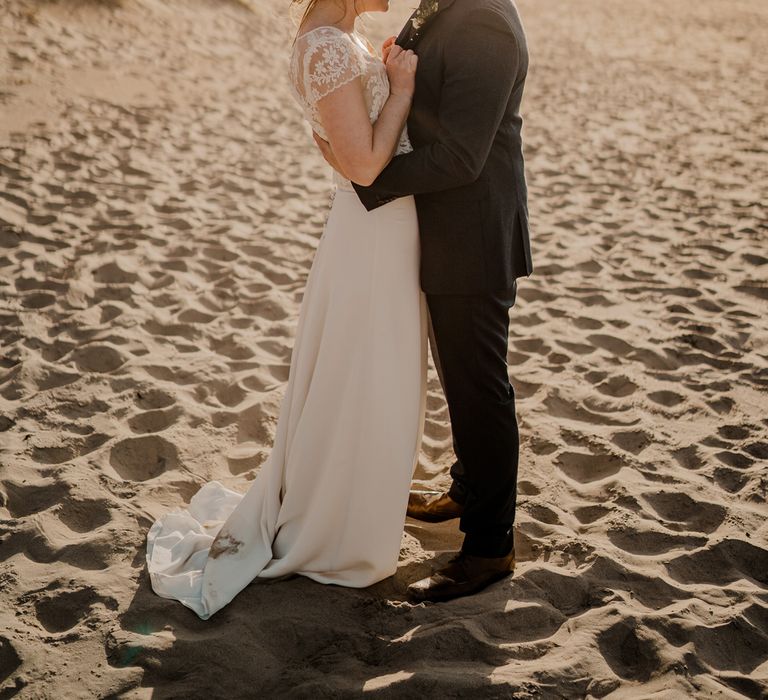 Groom in navy suit kisses forehead of bride in lace top capped sleeve wedding dress with satin skirt on the beach after Dunluce Castle wedding