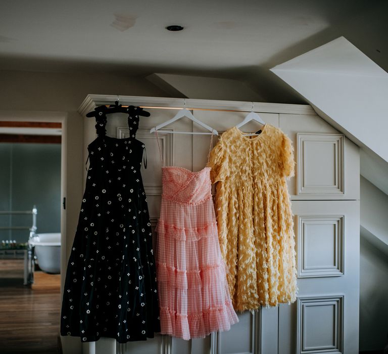 Long thick strapped printed black cami dress, midi pink ruffled dress and feathered yellow short sleeved dress all hanging on white wardrobe inside room at Hotel du Vin Harrogate for Harrogate wedding