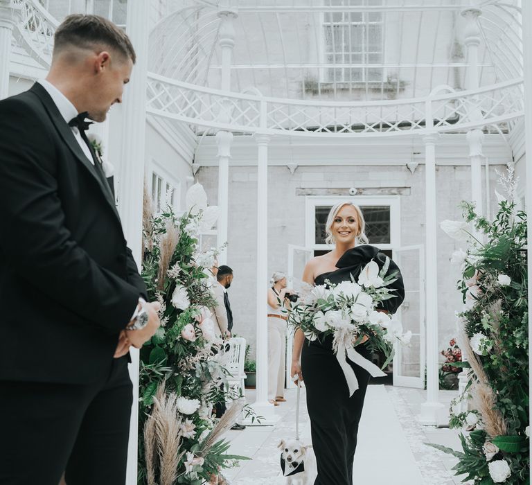 Maid of honour in black one-shoulder dress walks down the aisle in the glass conservatory at Came House Dorset holding white, pink and green bouquet and small white dog with tuxedo collar on a lead