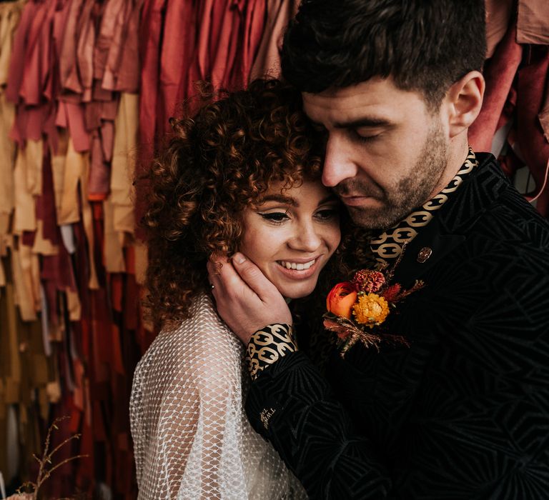 Groom in a black and gold patterned shirt with orange ranunculus buttonhole embracing his brides face with naturally curly hair