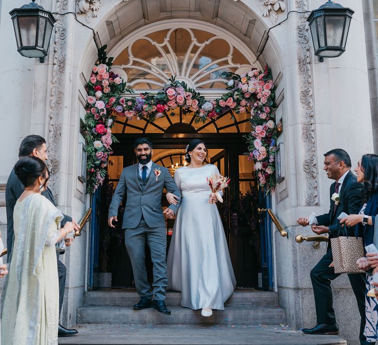 Bride & groom leave Chelsea Old Town Hall on their wedding day through a double door with brightly coloured rose archway complete with green foliage