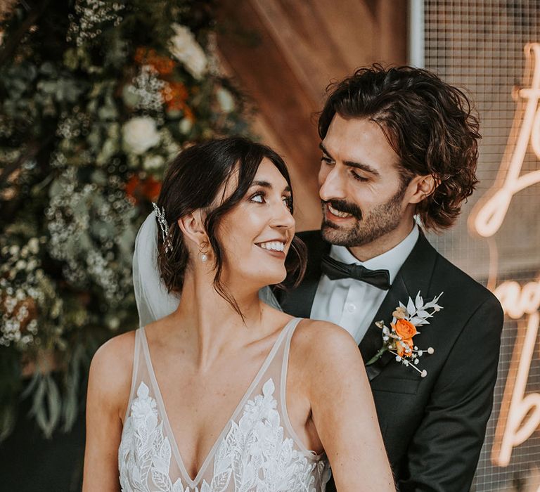 Groom in a black tuxedo with bow tie and orange buttonhole flower embracing his bride in an appliqué Made With Love wedding dress