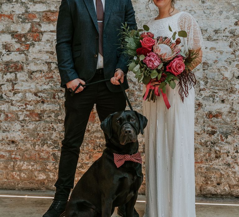 Bride & groom stand in front of brick wall with their dog wearing polka dot bow tie and bride holds brightly coloured floral bouquet