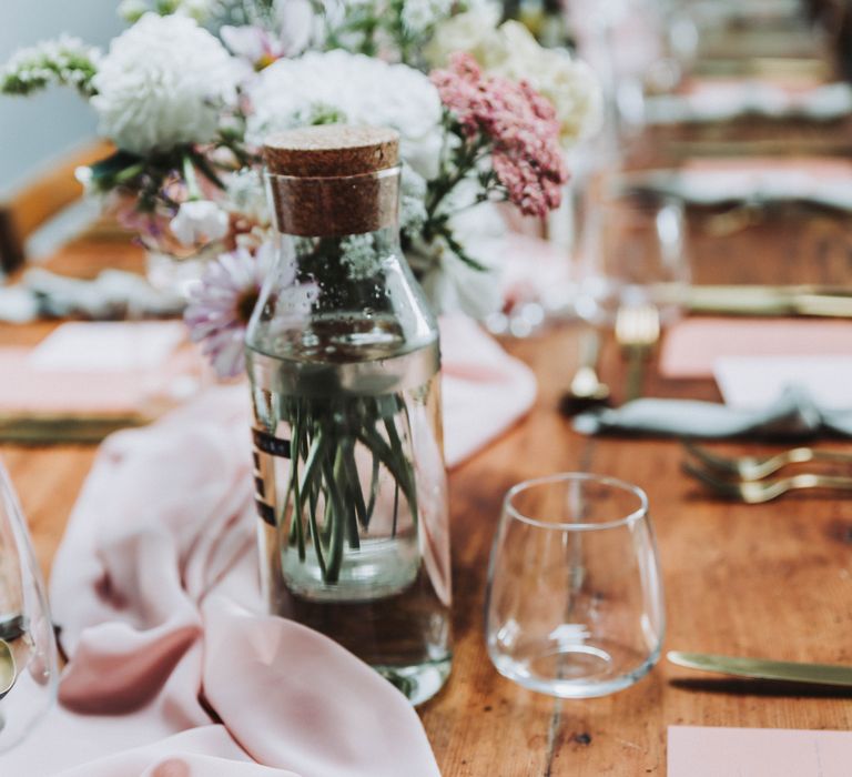 Pink table cloth runner across wooden table complete with glass water bottles with cork tops and gold cutlery 