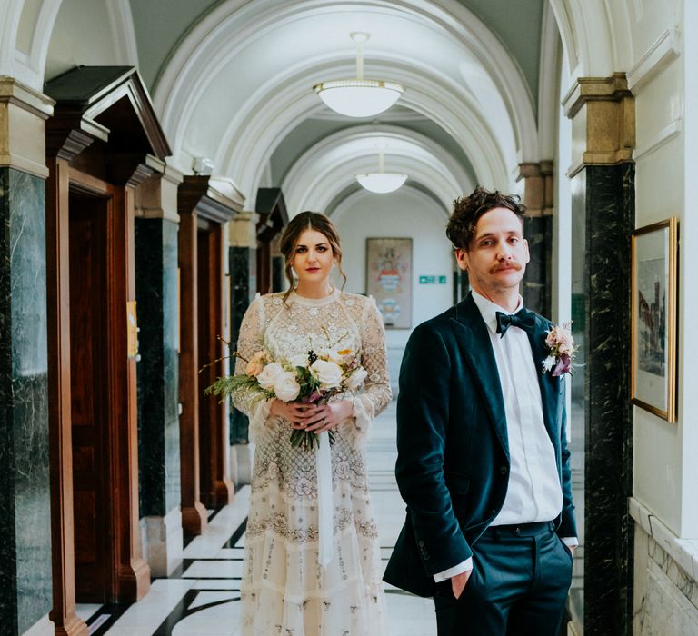 Bride & groom walk through Town Hall on their wedding day as groom look towards the camera whilst wearing velvet luxe suit