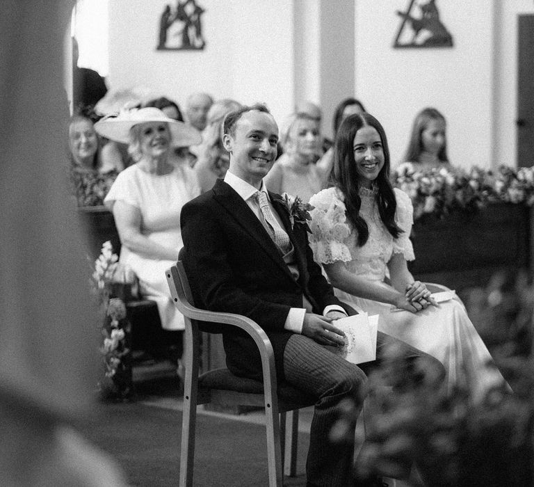 Bride in lace puffed sleeve Daalarna wedding dress smiles as she sits next to groom in black morning coat during church wedding ceremony in Surrey