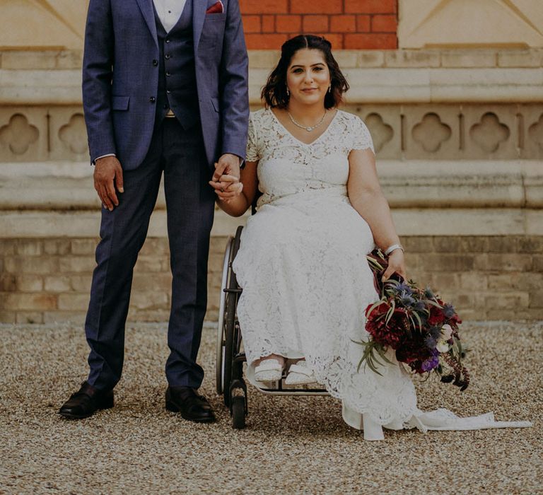 Groom in three-piece blue suit and burgundy bow tie holding hands with his bride in lace wedding dress as she sit in her wheelchair 