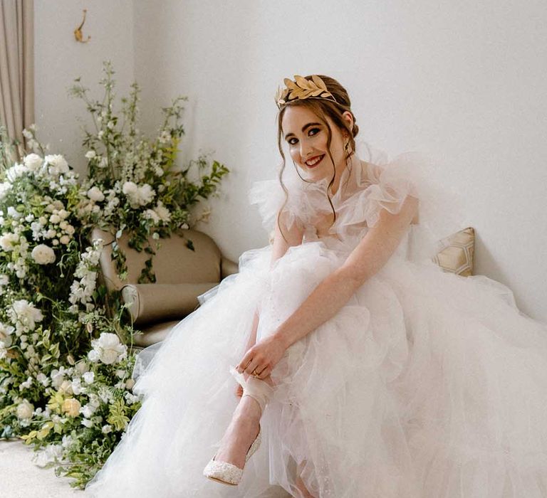 Bride in a tulle wedding dress with gold crown sitting on a chaise lounge putting on her embellished bridal shoes with ribbon ankle straps 