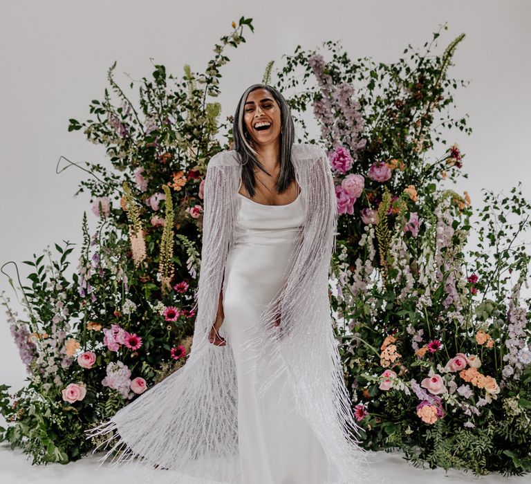 Bride in satin Halfpenny London wedding dress twirls tasselled Halfpenny London bridal cape whilst stood in front of multicoloured floral installation before wedding at Loft Studios London