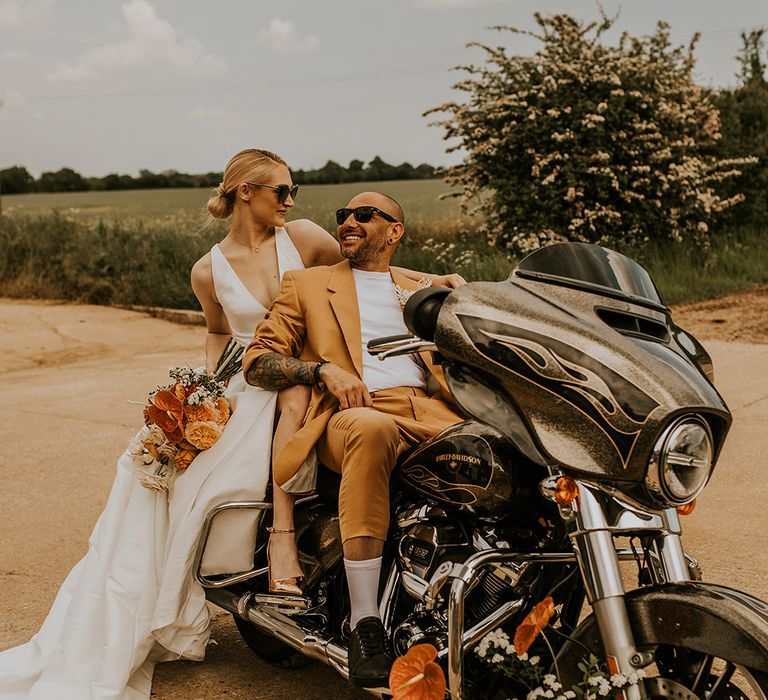 Stylish bride in a taffeta wedding dress with split design and groom in a mustard suit sitting on the back of their motorcycle wedding transport 