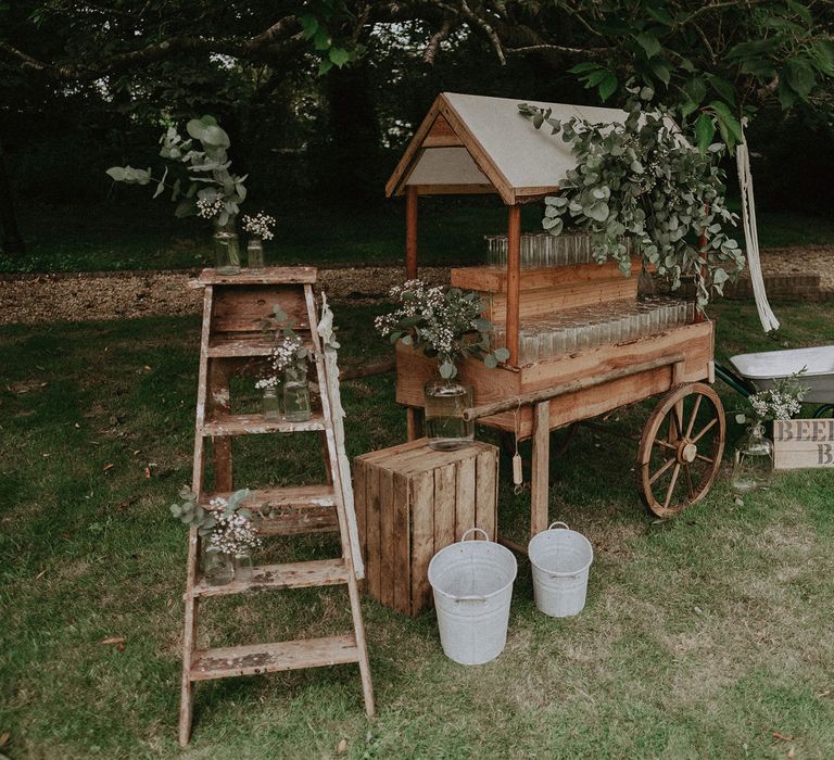 Outdoor bar cart with glasses, beer barrow and wooden boxes with milk churns at Isle of Wight wedding with macrame wedding decor