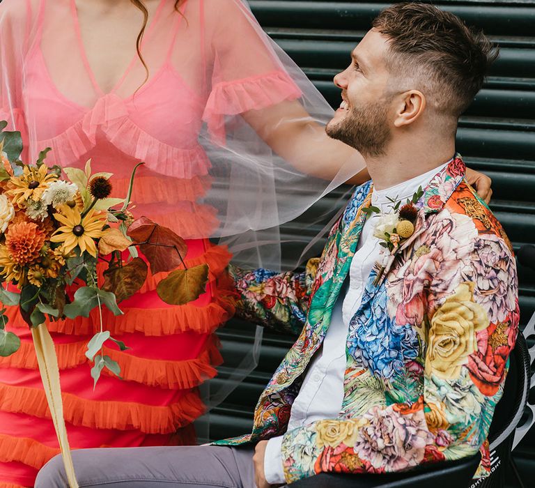 Bride in yellow dahlia flower crown with yellow earrings looking at her groom in a brightly coloured blazer 