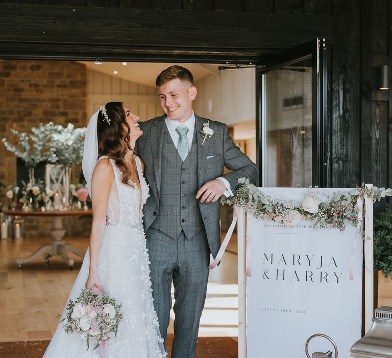 Bride in applique Berta wedding dress with headband and veil holds white, pink and green bridal bouquet as she stands with groom in grey checked suit next to white wedding welcome sign for summer wedding at Primrose Hill Farm 