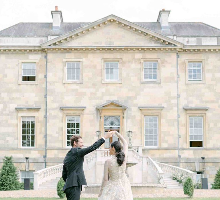 Groom in a tuxedo twirling his bride in a vintage lace wedding dress with long sleeves on the grounds of Botley's Mansion, Surrey 