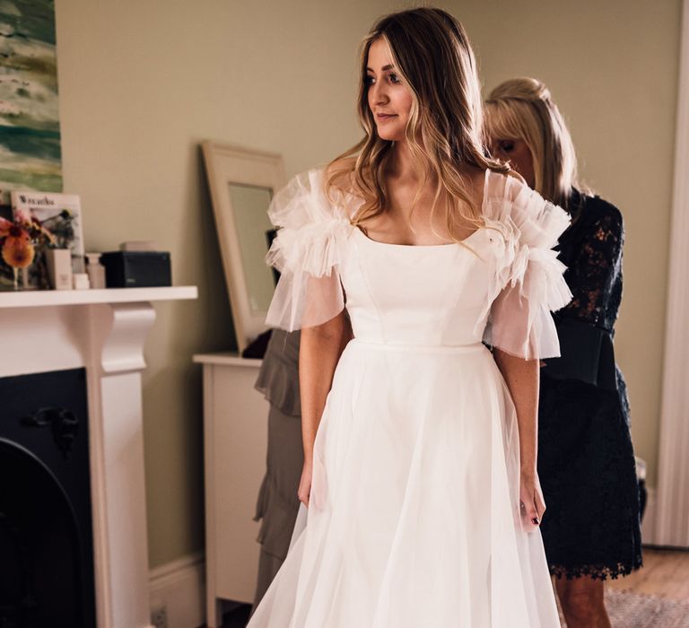 Bride in white Halfpenny London Mayfair dress with ruffle sleeves gets her dress zipped up by Wedding guests before church ceremony in Cornwall