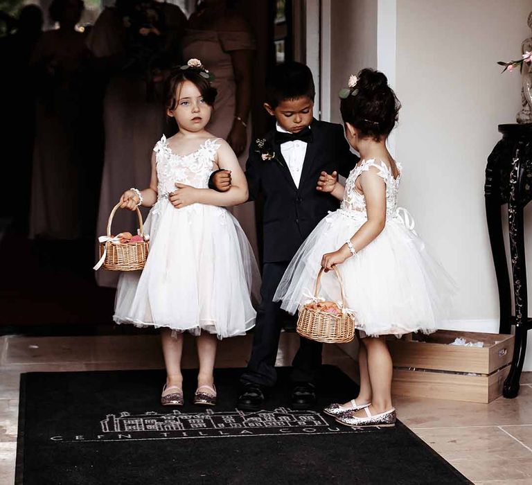 Flower girls in white dresses with sequins & wicker baskets hold hands with page boy in black suit with black bow tie 