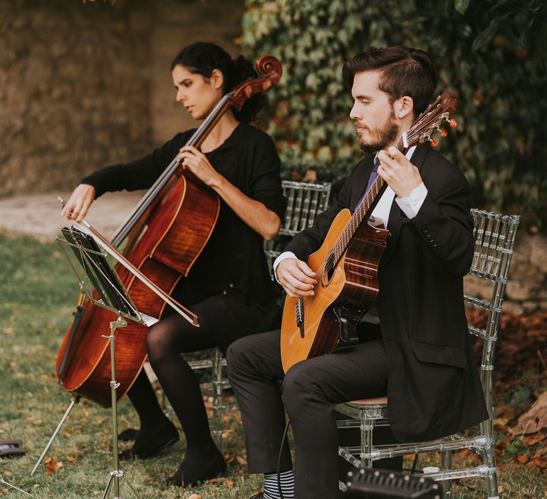 Guitar and cello player sit in perspex bamboo chairs playing outside during outdoor wedding ceremony at Notley Abbey