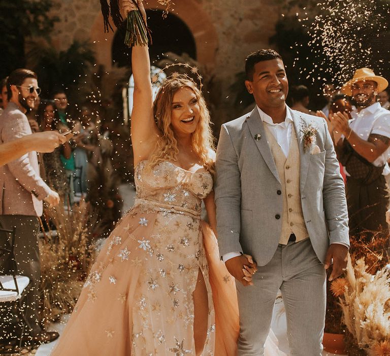 Bride & groom walk through confetti on their wedding day as bride lifts her bouquet in celebration