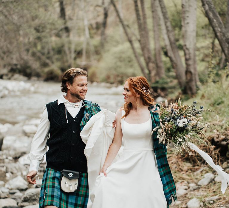 highland wedding inspiration with groom in a tartan kilt and bride in a princess wedding dress with plaid sash walking through a stream 