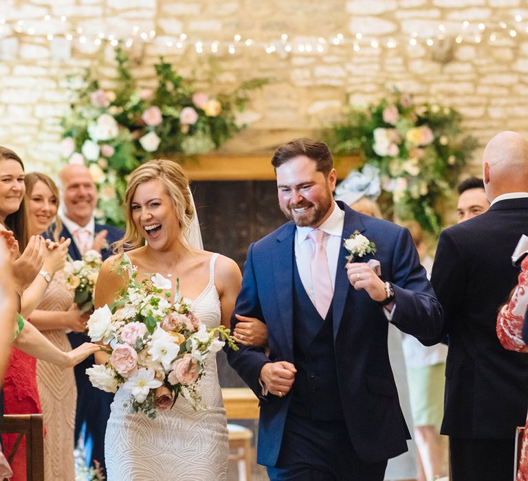 Bride in an embellished Made With Love wedding dress holding a pink and white bouquet descending up the aisle with her husband in a navy suit 