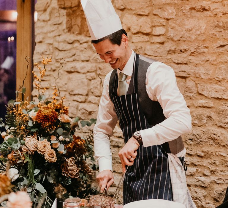 Smiling groom in striped apron and chefs hat cuts up meat at table during rustic barn wedding breakfast 