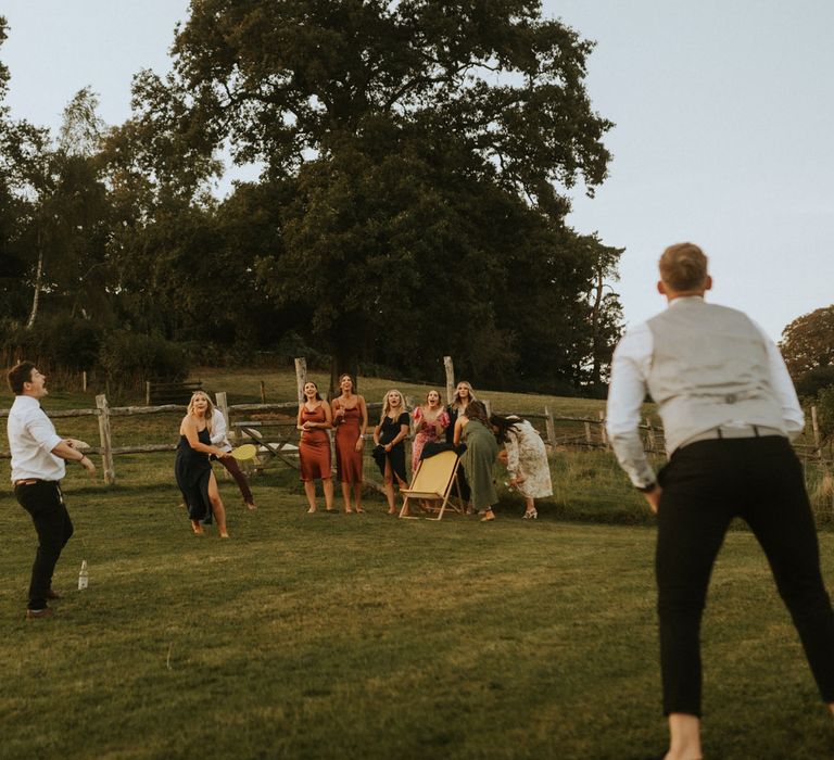 Wedding guests and bridesmaids in orange satin bridesmaid dresses play racquet sports during wedding at Hope Farm in Dorset