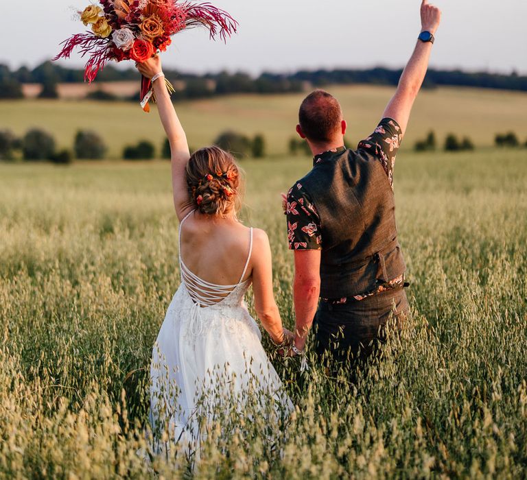 Bride in corset back wedding holding up rose and dried floral bouquet stands in field holding hands with groom in patterned short sleeve shirt after festival style wedding in Essex