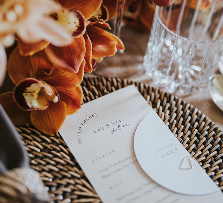 Stylish place setting with wicker place mat, menu card, champagne and orange wedding flowers 