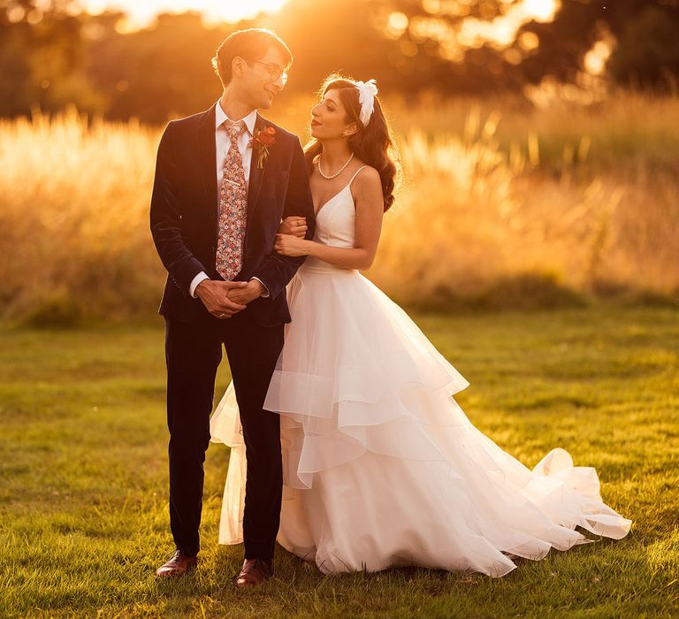 Bride in layered tulle wedding dress and headband stands holding arm of groom in dark suit and floral tie during golden hour after wedding at Elmore Court