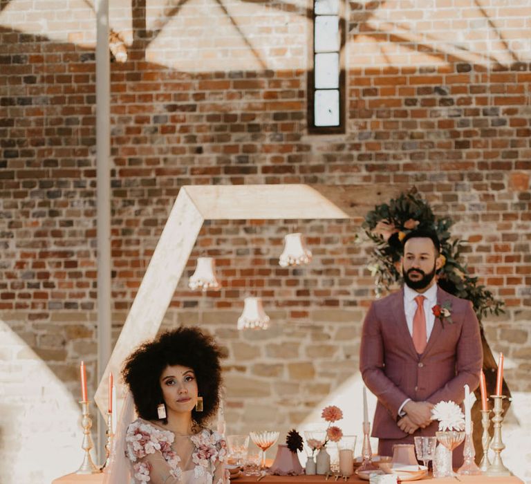 Black bride in a pink embroidered wedding dress sitting at an outdoor tables cape with orange tablecloth, pink lampshades and flower stems in vases 