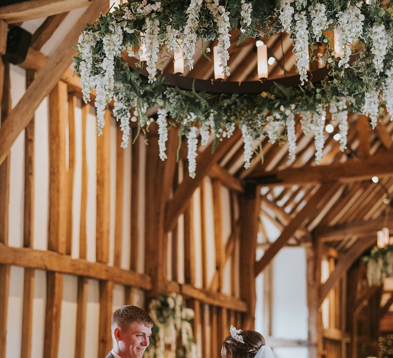 Groom in grey tweed suit and pink tie with bride at the altar with large rustic chandelier decorated with hanging wisteria 