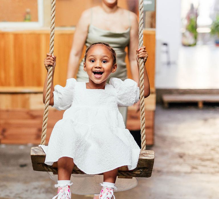 Smiling bright-eyed flower girl in white puff sleeve dress is swung on a swing by bridesmaids in sage green satin dress