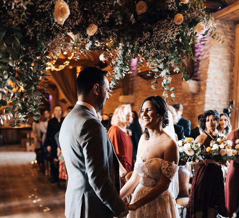 Bride smiles as she holds hands with the groom during their wedding ceremony under large white floral display