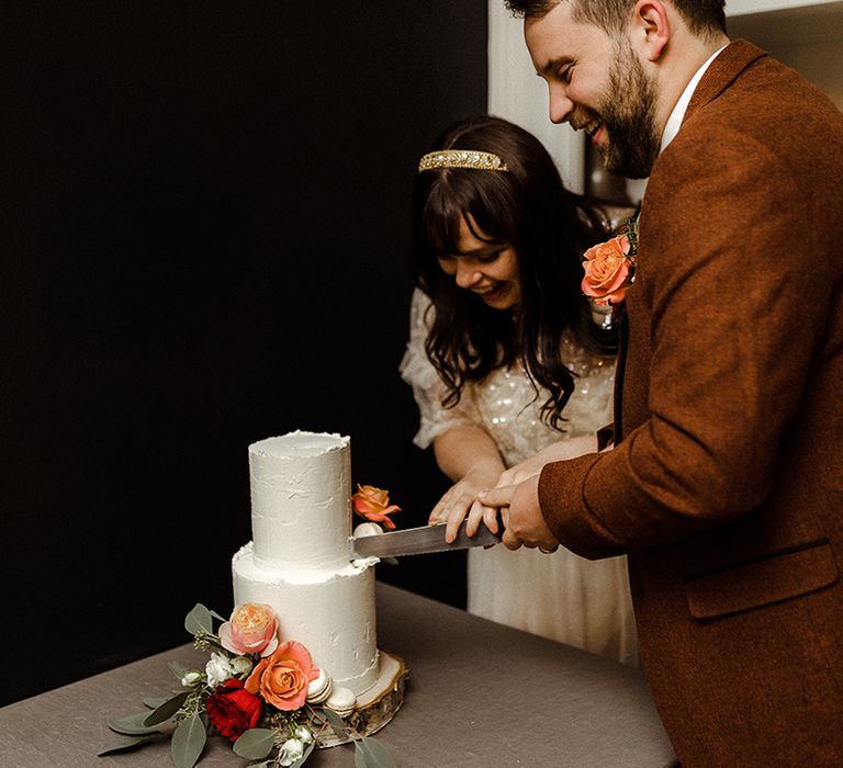 Bride and groom cut their two tier white buttercream wedding cake on a tree trunk stand decorated with roses and macaroons