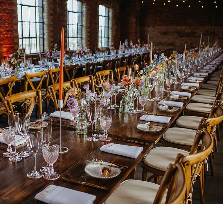 Table setting with long banquet tables decorated with colourful spring flowers, orange and cream candles and pink and green coloured glassware