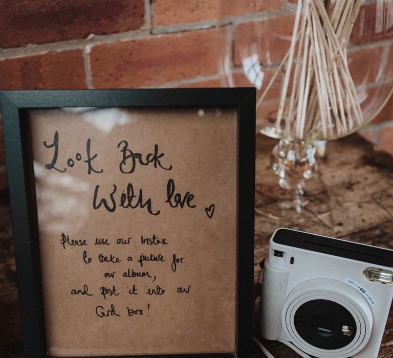 Handwritten sign in black photo frame requesting guests take a picture of themselves with polaroid camera and place it in the guest book