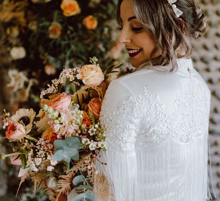 Modern bride in a satin wedding dress with embroidered shoulder and tassel detail earring a crystal headband and holding a blush wedding bouquet 