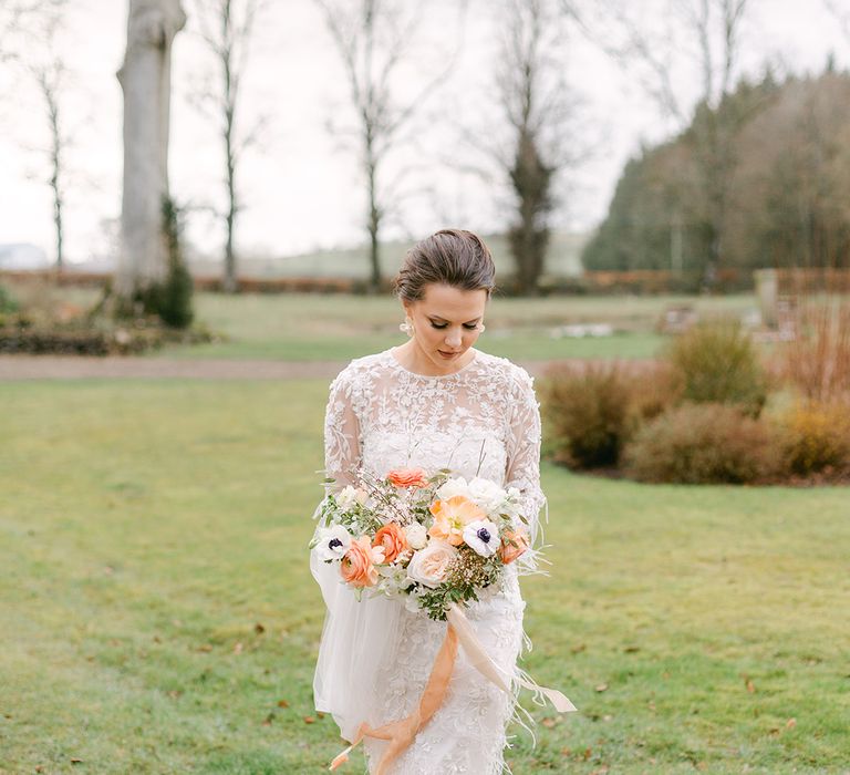 Bride in a midi applique and feather wedding dress with long sleeves holding a peach and white wedding bouquet 